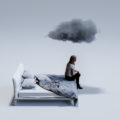 Tired mid adult woman sitting on corner of bed with dark cloud above head.