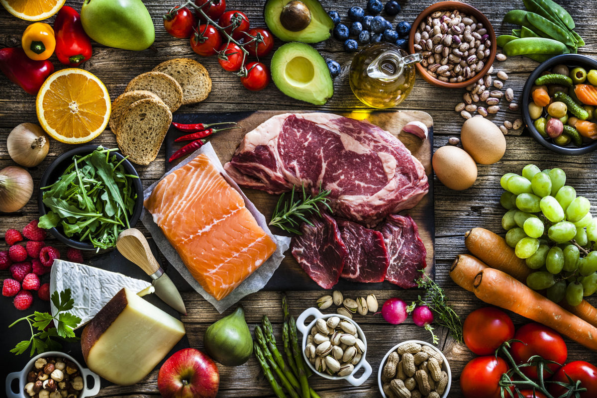 Food backgrounds: top view of a rustic wooden table filled with different types of food. At the center of the frame is a cutting board with beef steak and a salmon fillet and all around it is a large variety of food like fruits, vegetables, cheese, bread, eggs, legumes, olive oil and nuts. DSRL studio photo taken with Canon EOS 5D Mk II and Canon EF 70-200mm f/2.8L IS II USM Telephoto Zoom Lens