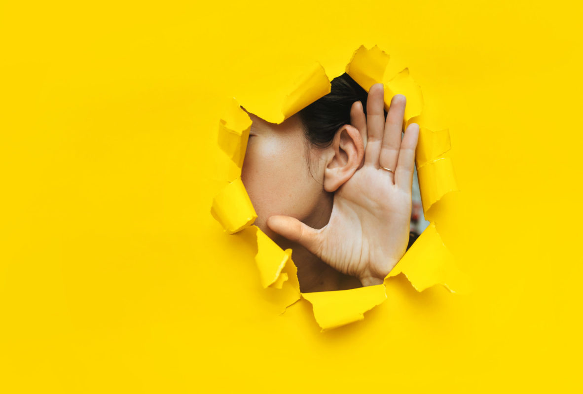 Close-up of a woman's ear and hand through a torn hole in yellow paper.