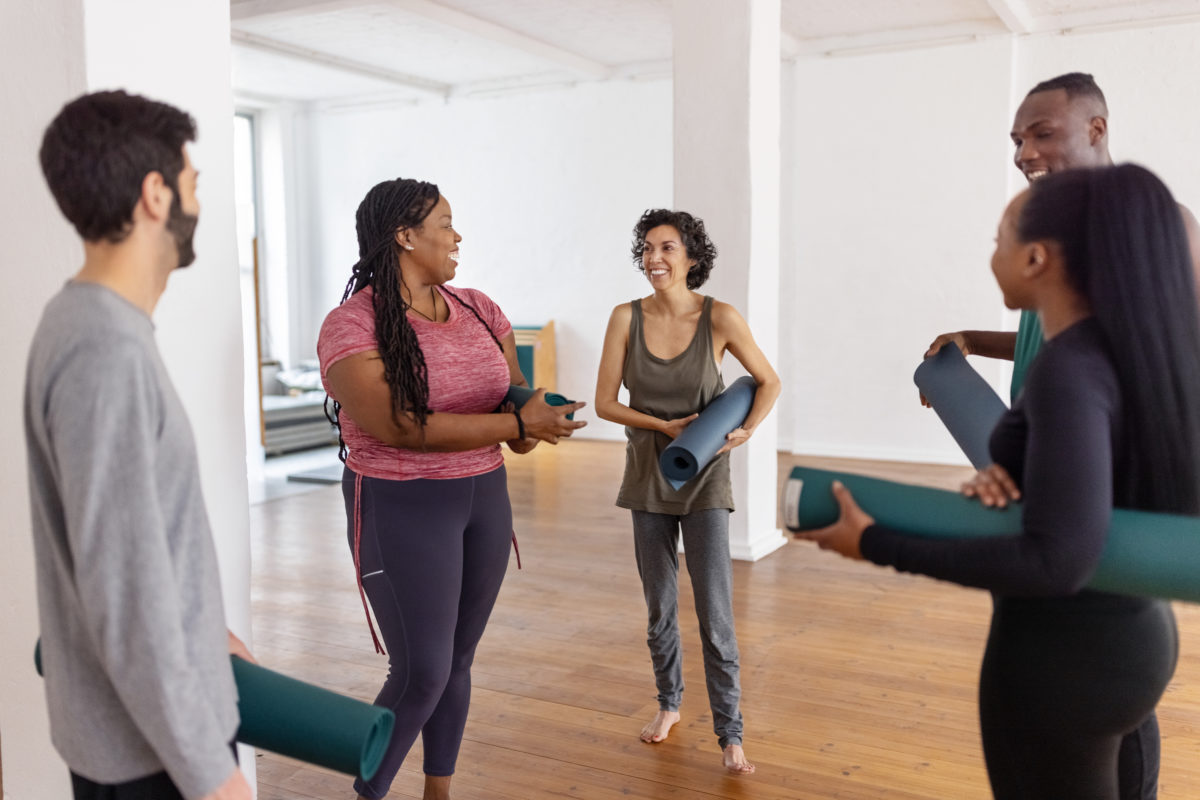 Group of diverse people holding yoga mats talking after a yoga class. Multiracial group of fit people talking and relaxing in sports club after workout training.
