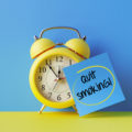 Yellow alarm clock with a blue post it note attached over bright blue background. Quit smoking writes on post it note. Reminder concept. Horizontal composition with copy space.