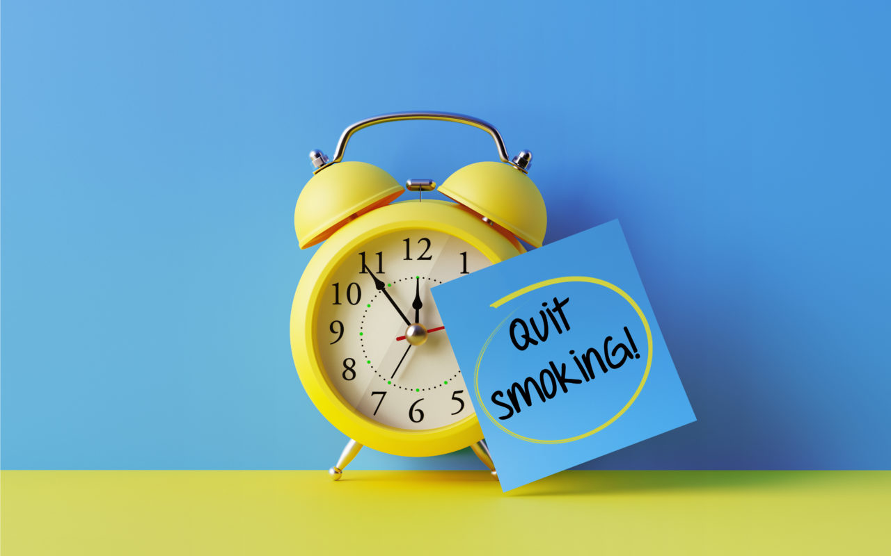Yellow alarm clock with a blue post it note attached over bright blue background. Quit smoking writes on post it note. Reminder concept. Horizontal composition with copy space.