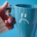 Blue Monday concept. Close-up of a female hand in a white sweater holds a blue cup with a sad smiley as a symbol of the most depressive day of the year on a blue background