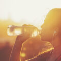 Silhouette of a woman drinking form a water bottle. She is exercising at sunset or sunrise. She has a mobile phone on her arm and is listening to music.