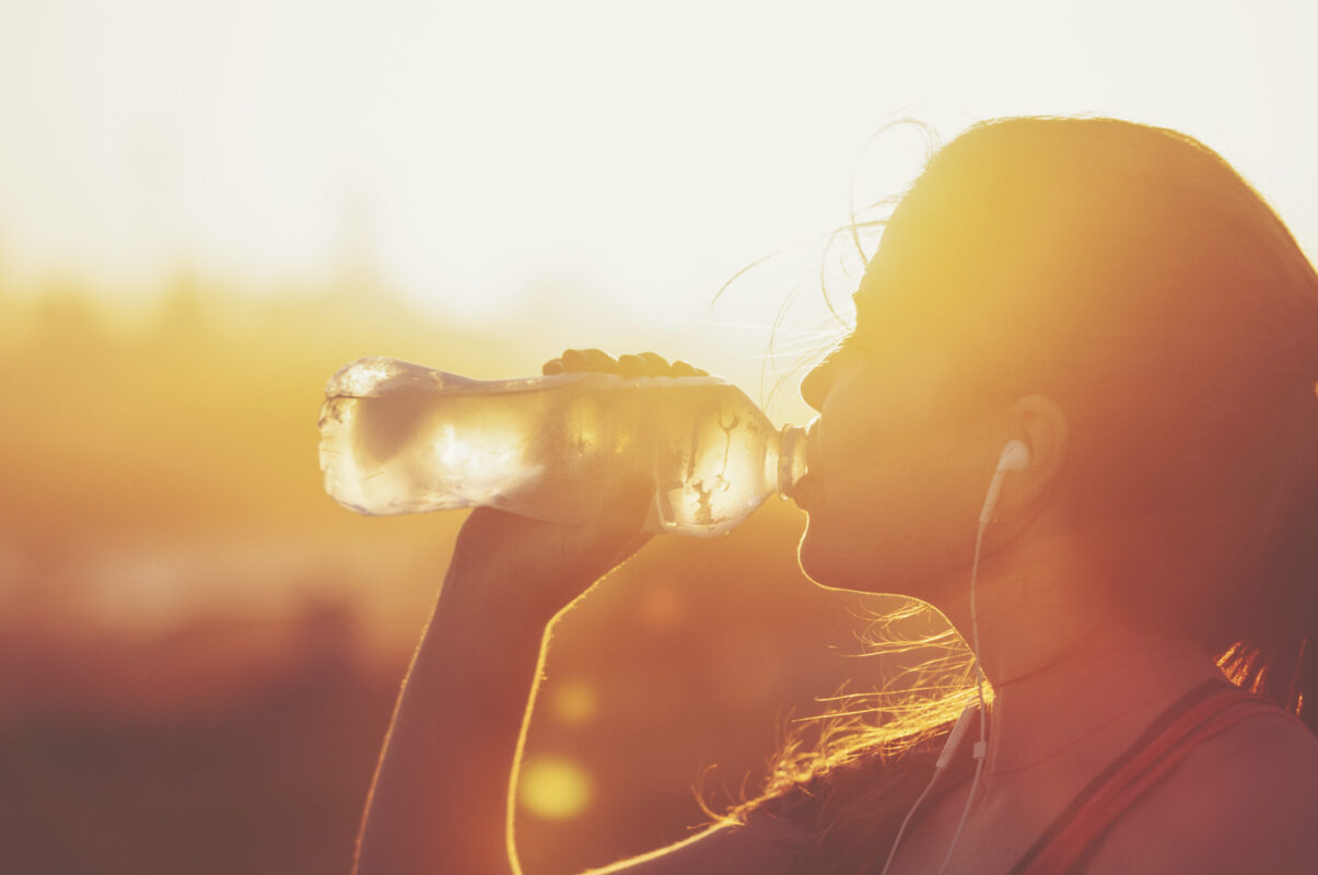 Silhouette of a woman drinking form a water bottle. She is exercising at sunset or sunrise. She has a mobile phone on her arm and is listening to music.