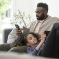 A man and daughter recline on the couch whilst giving their attention to their phone and tablet