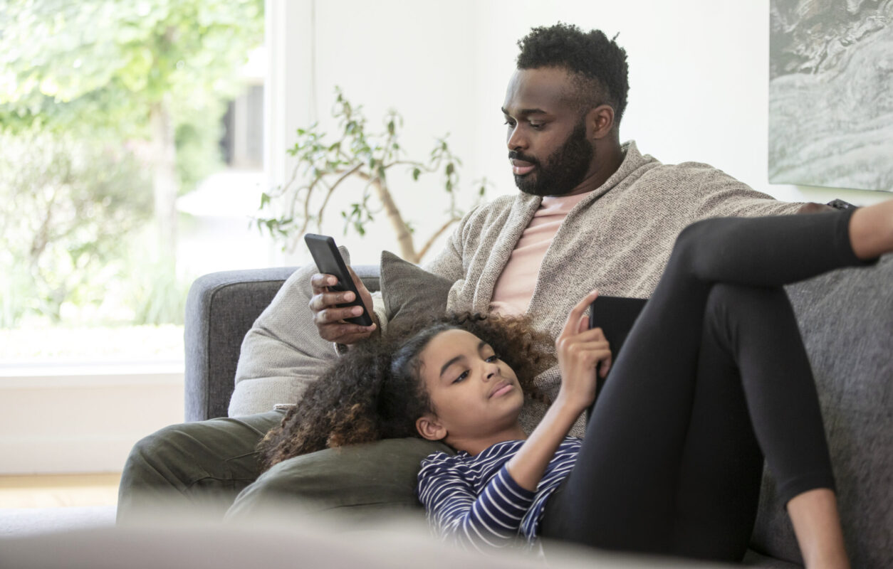A man and daughter recline on the couch whilst giving their attention to their phone and tablet