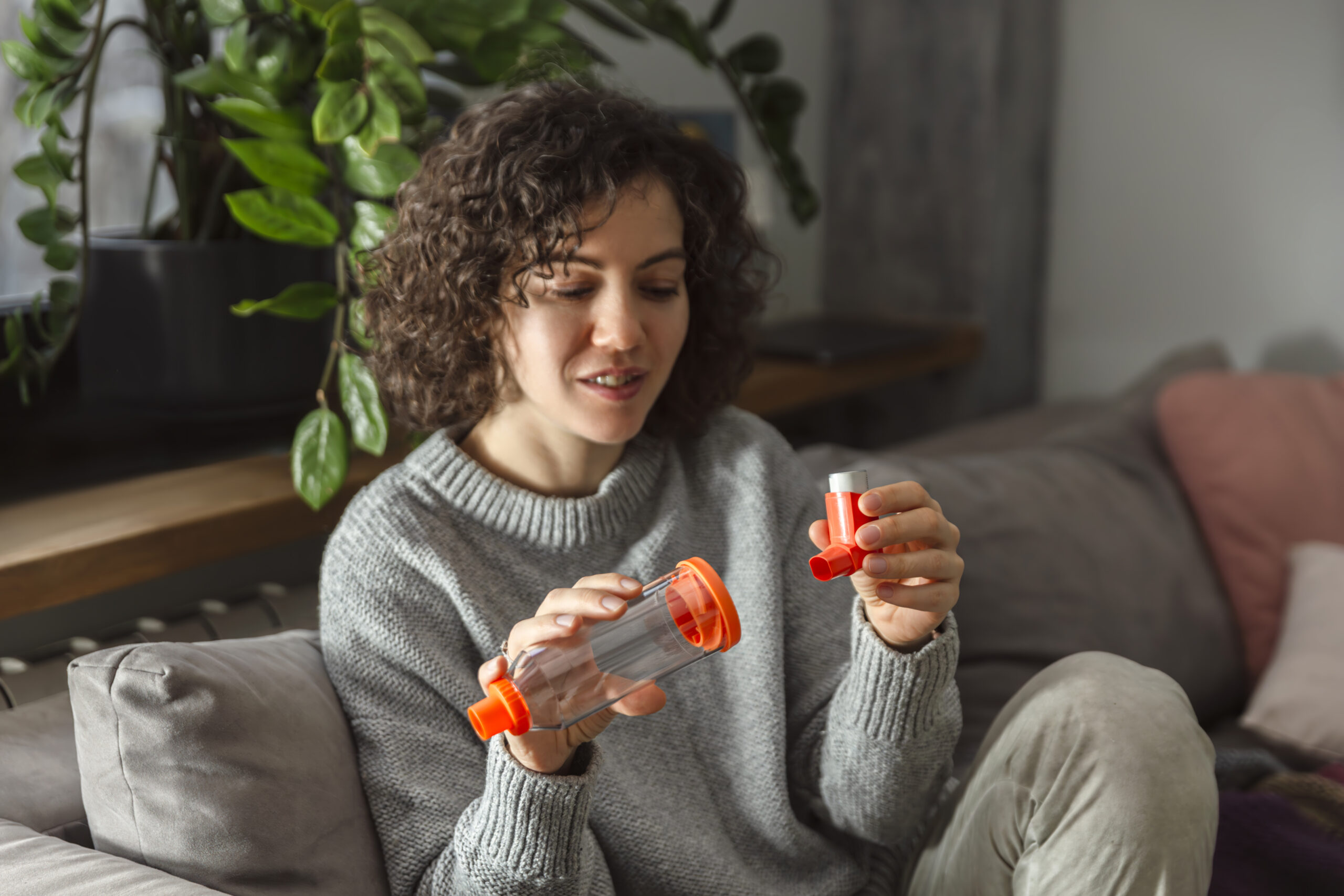 Young adult woman takes her asthma medication with an inhaler while sitting on a sofa in living room