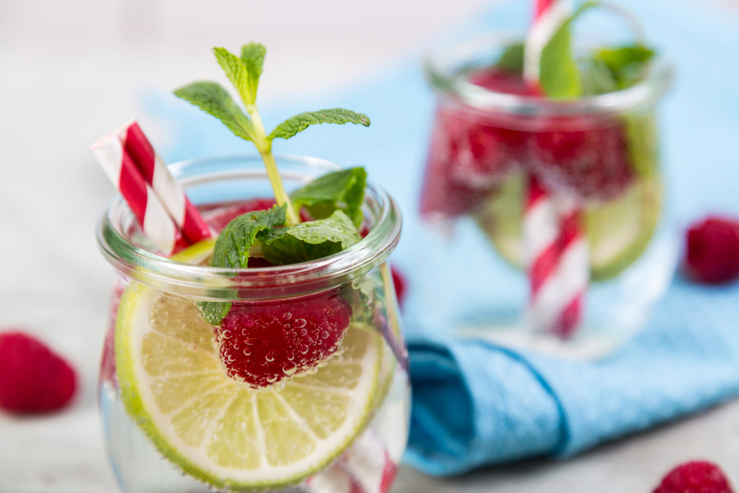Little glass jars with fresh mint, lemon slice and raspberries refresher concept, over white wooden table with blue napkin
