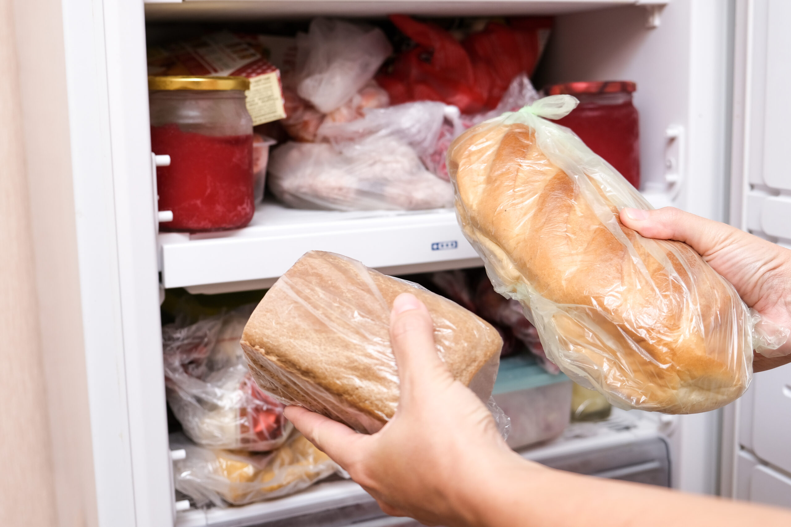 A hand putting two loafs of wheat and brown bread in reserve on a shelf of a home freezer.