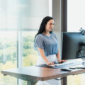 Using a standing desk, the adult female works on her computer in her office.