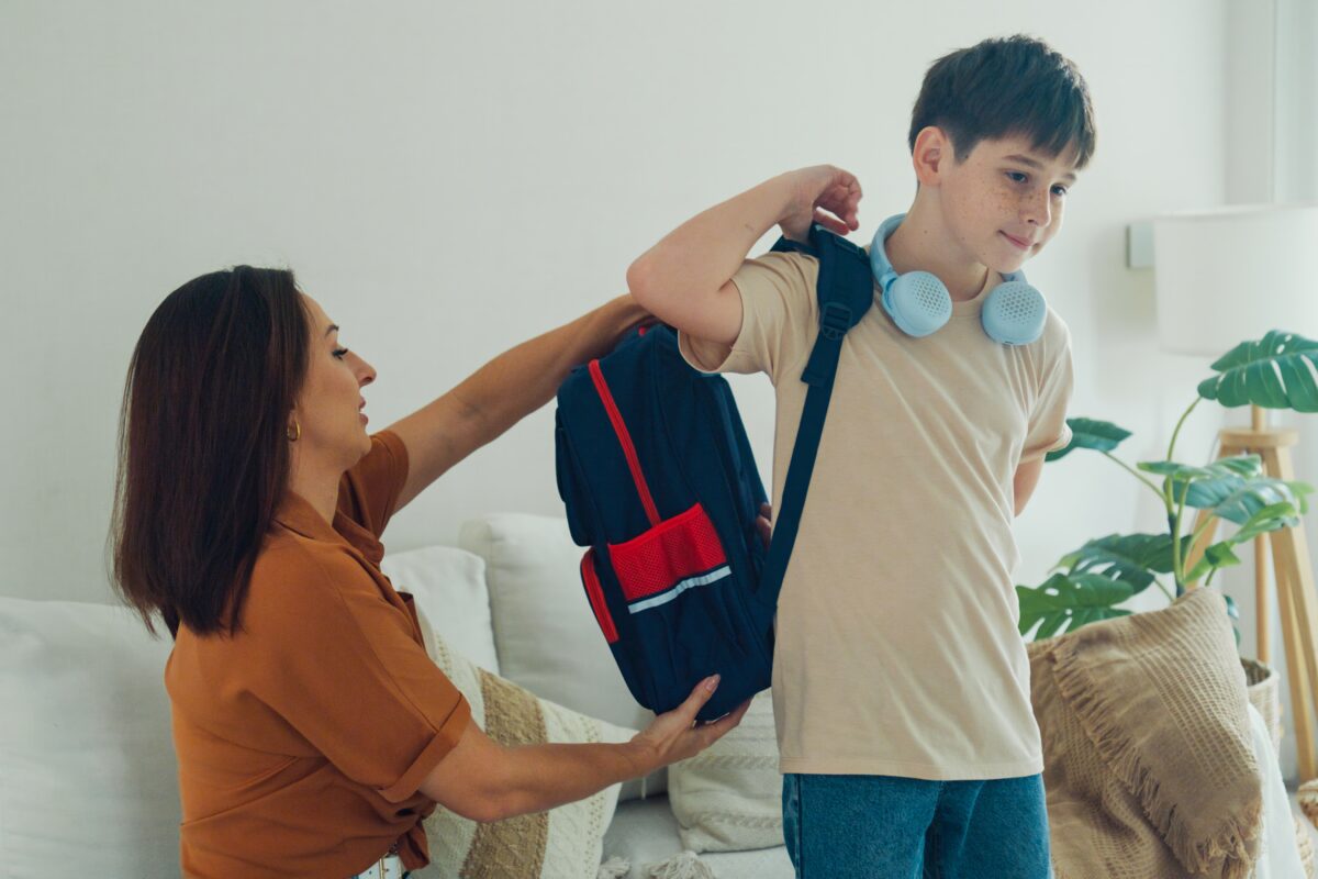 Young mother helps her son prepare his backpack in the living room for school
